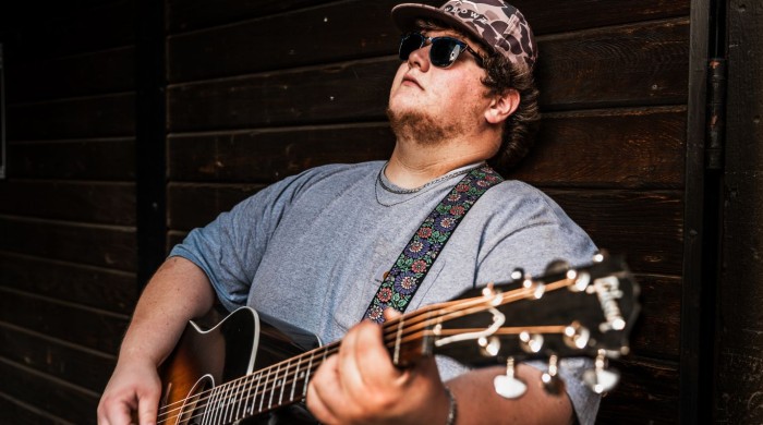 A man leaning against a wooden wall holding a guitar wearing a blue tshirt, camo hat, and black sunglasses